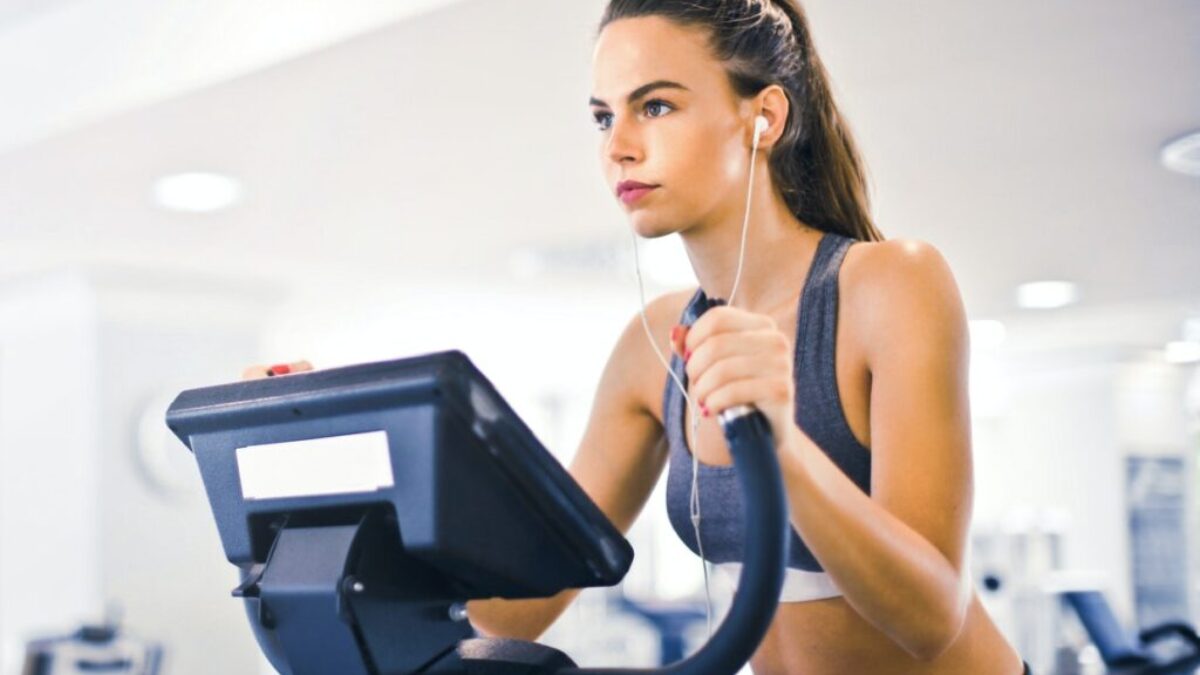 Serious fit woman in earphones and activewear listening to music and running on treadmill in light contemporary sports center