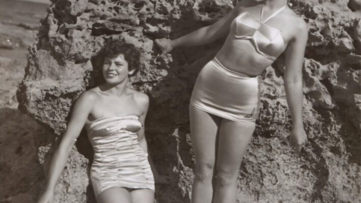 grayscale photo of three women standing and sitting on rocks
