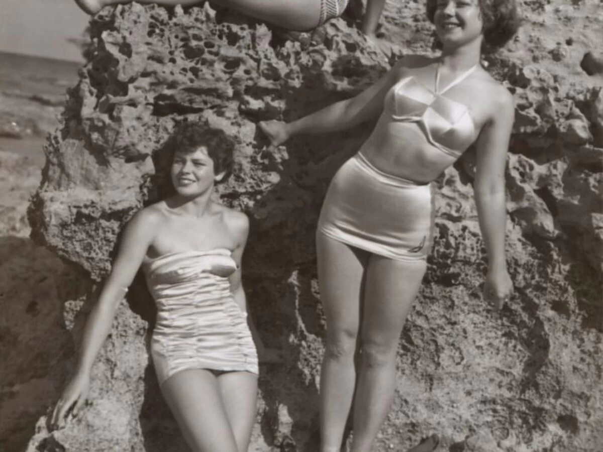grayscale photo of three women standing and sitting on rocks