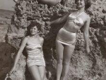grayscale photo of three women standing and sitting on rocks