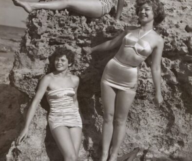 grayscale photo of three women standing and sitting on rocks
