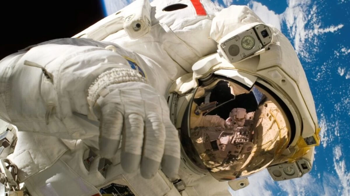 This picture shows an american astronaut in his space and extravehicular activity suite working outside of a spacecraft. In the background parts of a space shuttle are visible. In the far background of the picture planet earth with it's blue color and white clouds is shown as well as a patch of black space.