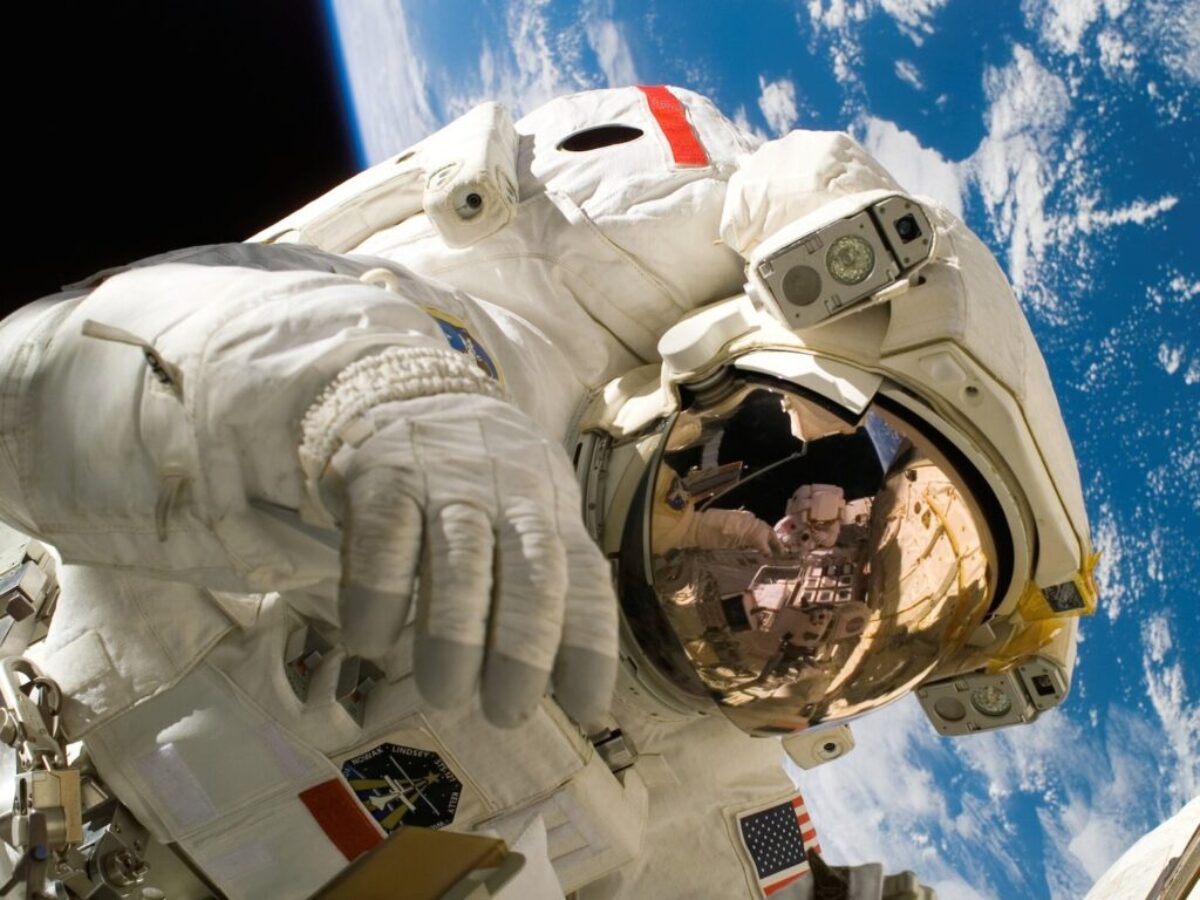 This picture shows an american astronaut in his space and extravehicular activity suite working outside of a spacecraft. In the background parts of a space shuttle are visible. In the far background of the picture planet earth with it's blue color and white clouds is shown as well as a patch of black space.