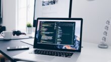 A MacBook with lines of code on its screen on a busy desk