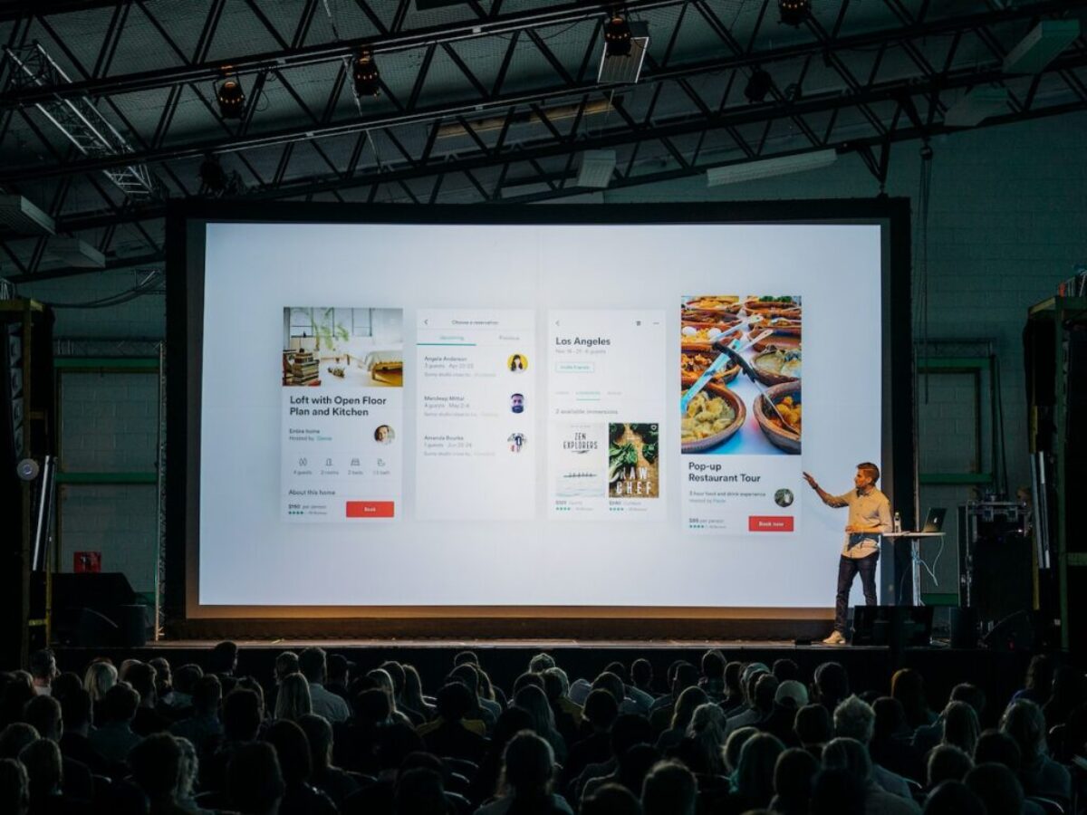 person discussing while standing in front of a large screen in front of people inside dim-lighted room