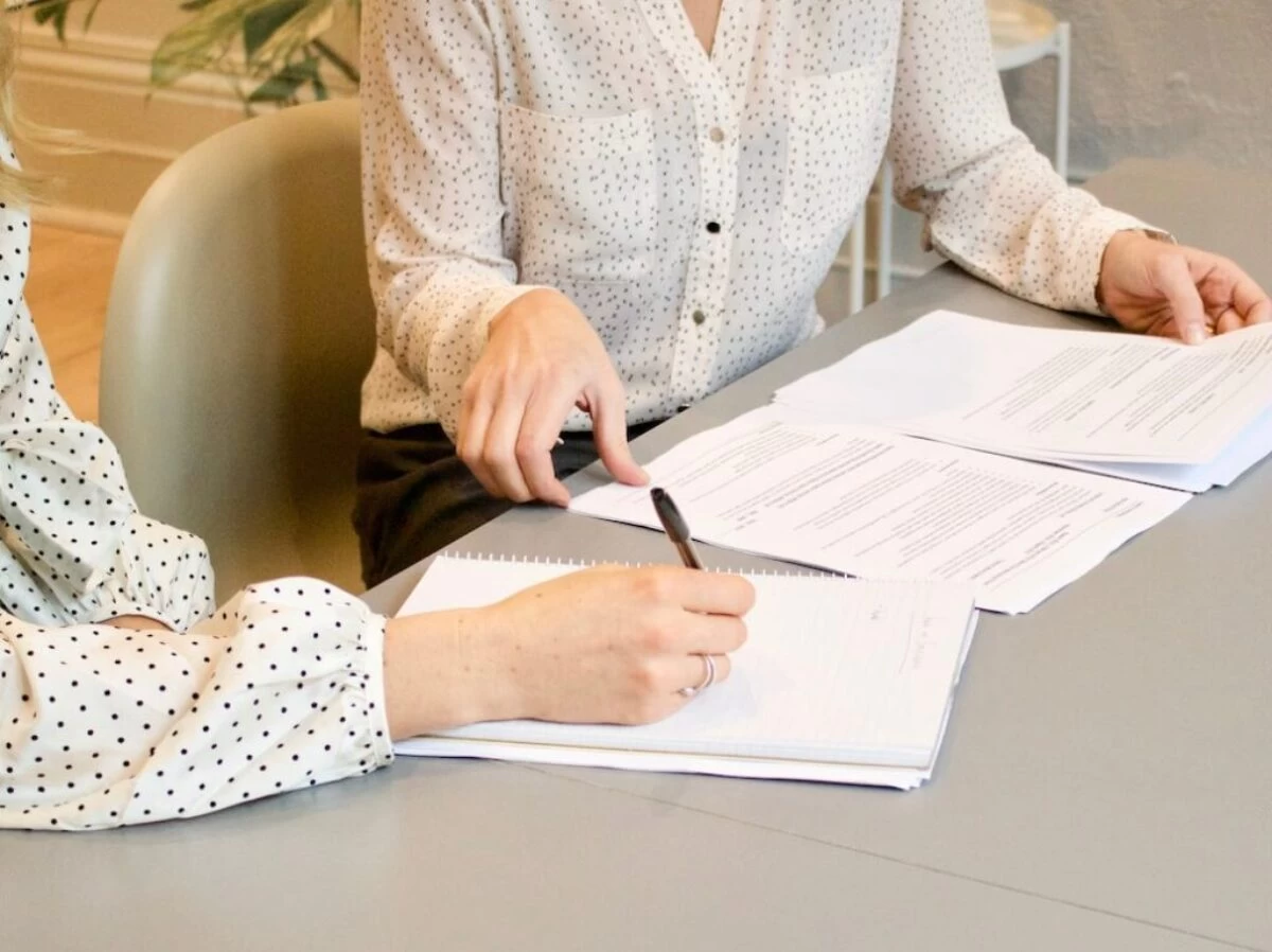 woman signing on white printer paper beside woman about to touch the documents