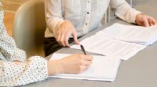 woman signing on white printer paper beside woman about to touch the documents