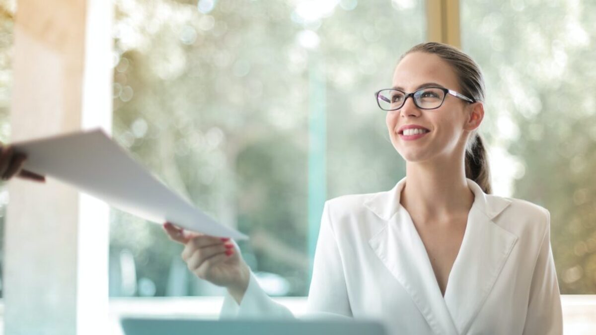 Low angle of successful female executive manager in classy style sitting at table with laptop in contemporary workplace and passing documents to colleague