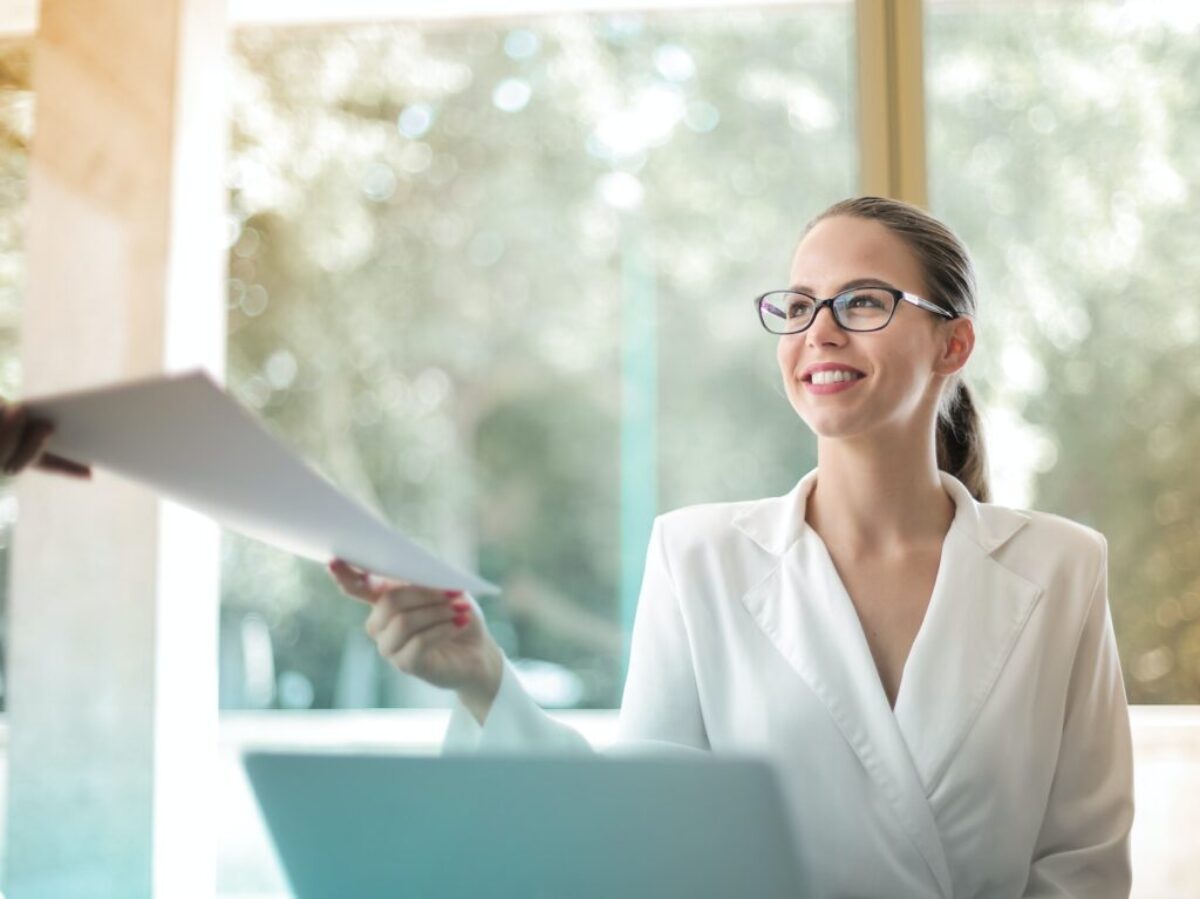 Low angle of successful female executive manager in classy style sitting at table with laptop in contemporary workplace and passing documents to colleague