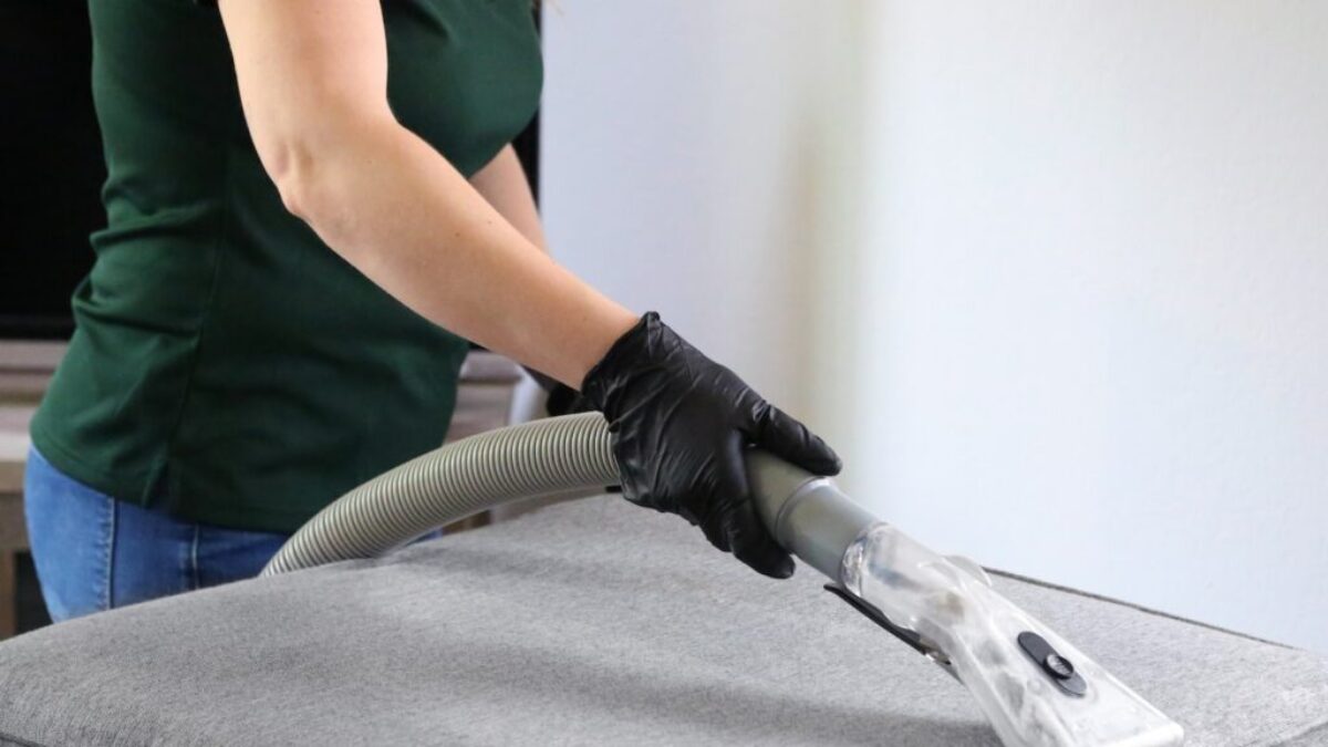 a woman in a green shirt and black gloves vacuuming a gray ottoman