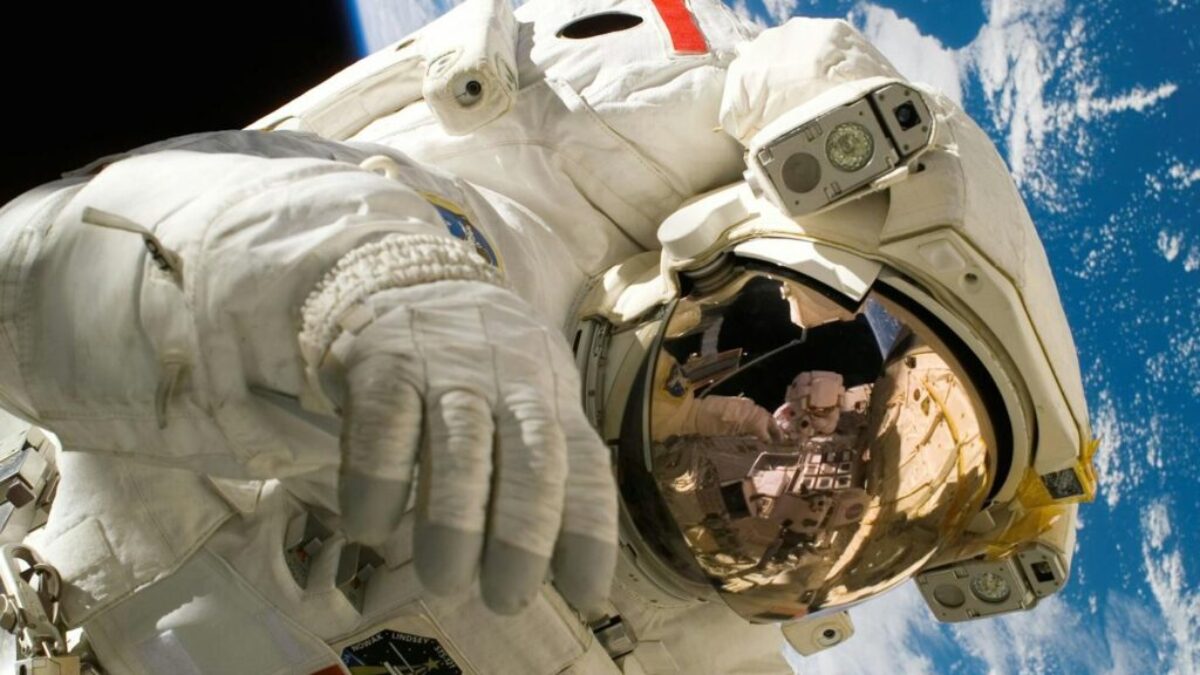 This picture shows an american astronaut in his space and extravehicular activity suite working outside of a spacecraft. In the background parts of a space shuttle are visible. In the far background of the picture planet earth with it's blue color and white clouds is shown as well as a patch of black space.
