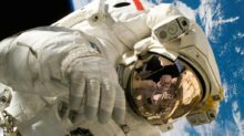 This picture shows an american astronaut in his space and extravehicular activity suite working outside of a spacecraft. In the background parts of a space shuttle are visible. In the far background of the picture planet earth with it's blue color and white clouds is shown as well as a patch of black space.