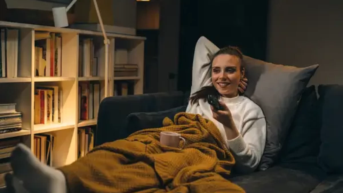 Žena pod dekou na gauči sleduje televizi