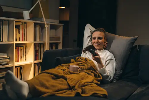 Žena pod dekou na gauči sleduje televizi