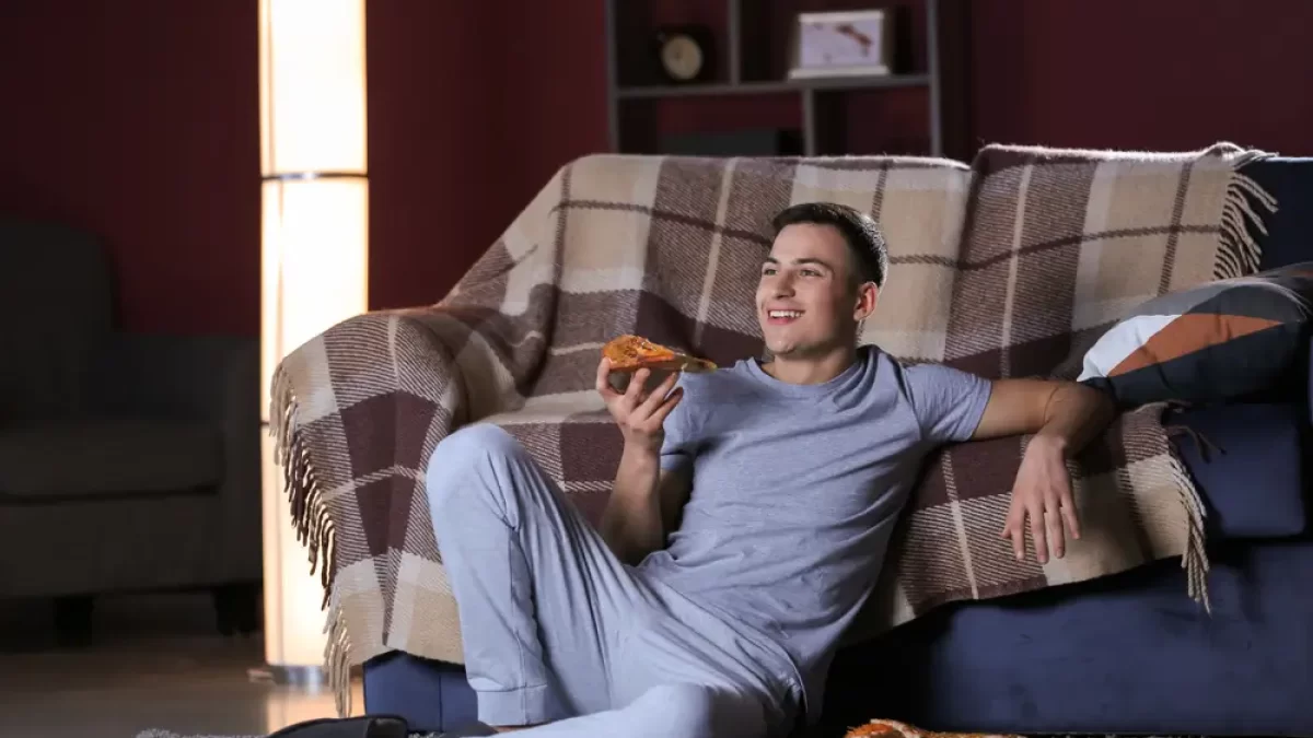 Muž se v noci dívá na televizi a jí u toho pizzu