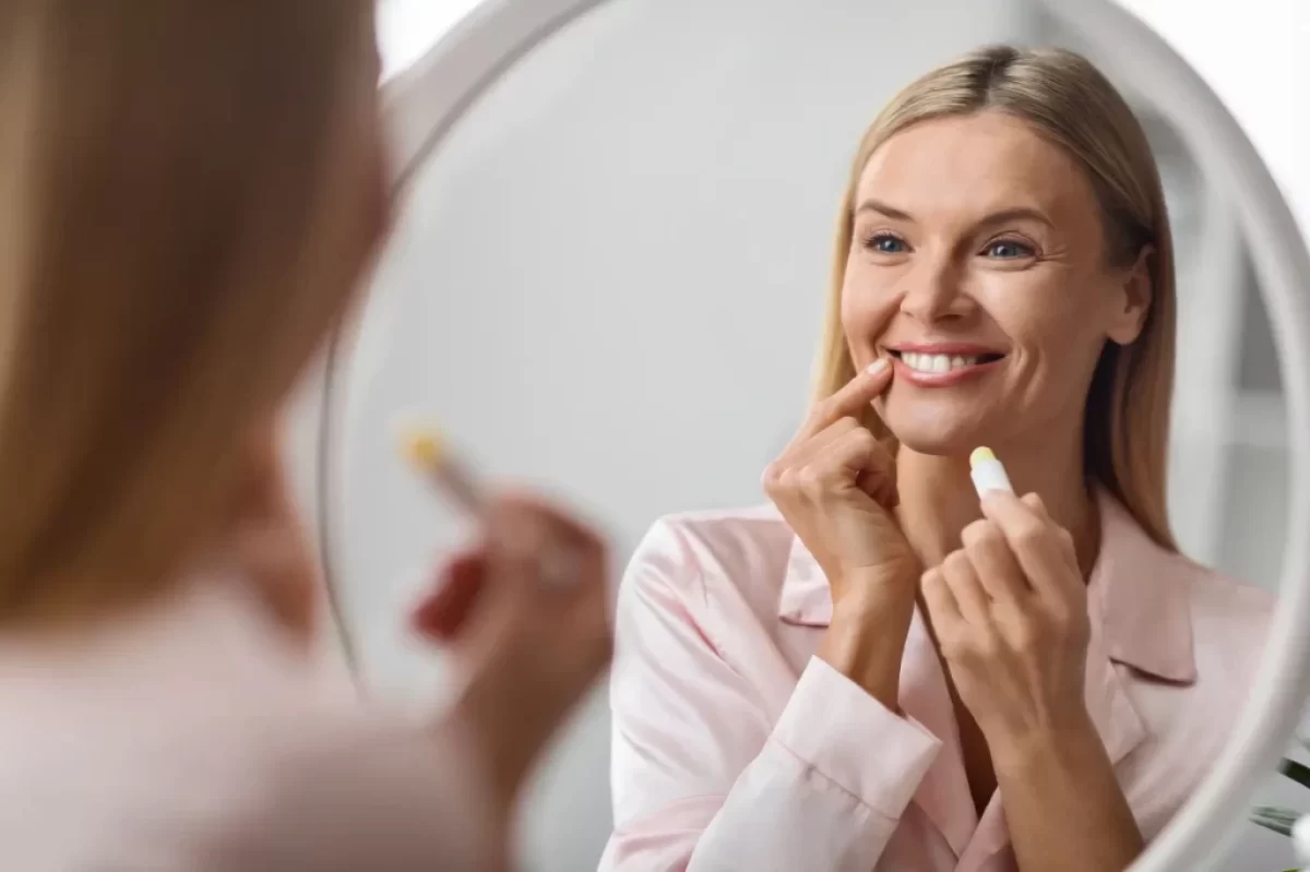Žena se na sebe dívá do zrcadla a aplikuje balzám na rty