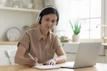 Žena s notebookem a sluchátky píše do sešitu