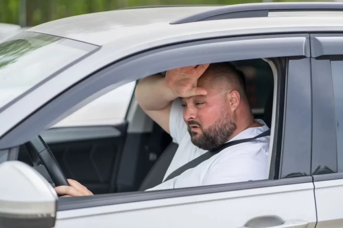 Muž v bílém tričku za volantem si otírá pot z čela.
