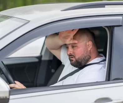 Muž v bílém tričku za volantem si otírá pot z čela.