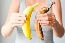 Žena ve světlém tílku drží v každé ruce jeden banán. V levé ruce je čerstvý, jasně žlutý banán, zatímco v pravé ruce je banán výrazně přezrálý, hnědý a skvrnitý.