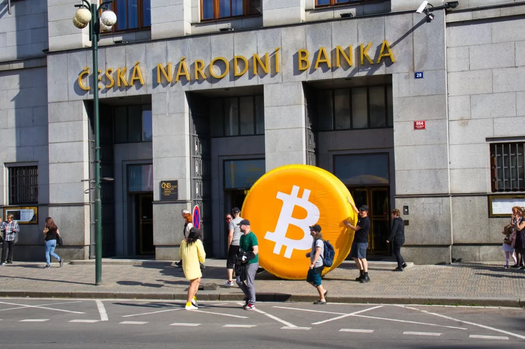 bit3 - Techsvět Fotografie zachycuje skupinu lidí, kteří nesou obří nafukovací symbol Bitcoinu před budovu České národní banky v Praze.