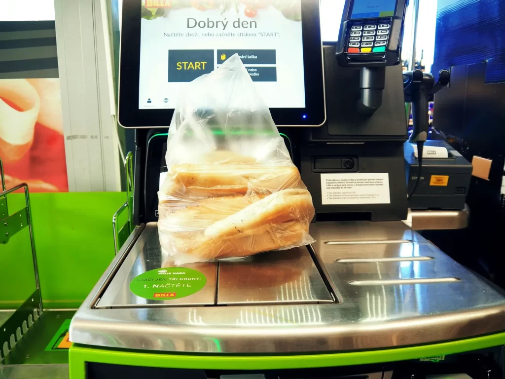 black2 - Techsvět Na obrázku je samoobslužná pokladna v supermarketu Billa. Na váhové ploše pokladny leží igelitový sáček s několika rohlíky. Displej pokladny zobrazuje výzvu „Dobrý den – načtěte zboží nebo stiskněte START.“