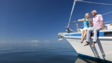 Starší pár sedí na přídi jachty. Oba se usmívají a užívají si slunečný den na klidném moři pod modrou oblohou.