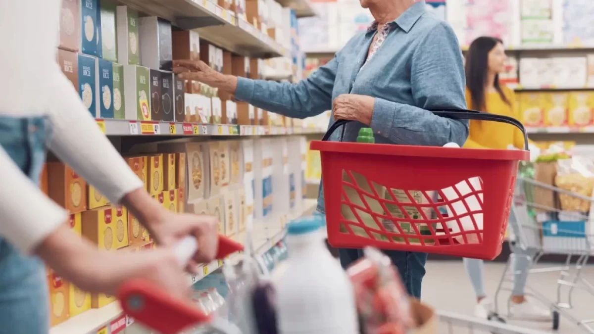 Žena nakupující v supermarketu. V levé ruce drží červený nákupní košík, pravou sahá po zboží na horní polici regálu.