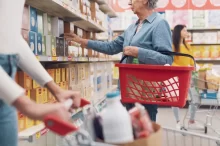 Žena nakupující v supermarketu. V levé ruce drží červený nákupní košík, pravou sahá po zboží na horní polici regálu.