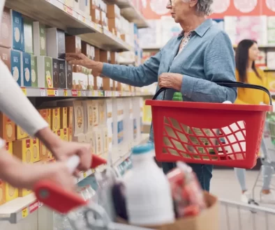Žena nakupující v supermarketu. V levé ruce drží červený nákupní košík, pravou sahá po zboží na horní polici regálu.