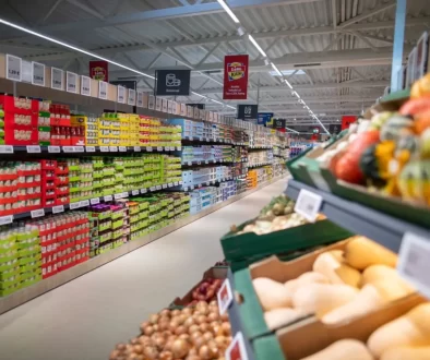 Interiér prodejny Lidl. Vlevo vidíme dlouhou řadu regálů plných trvanlivých potravin, jako jsou konzervy a balené výrobky, uspořádané podle barev a typů.