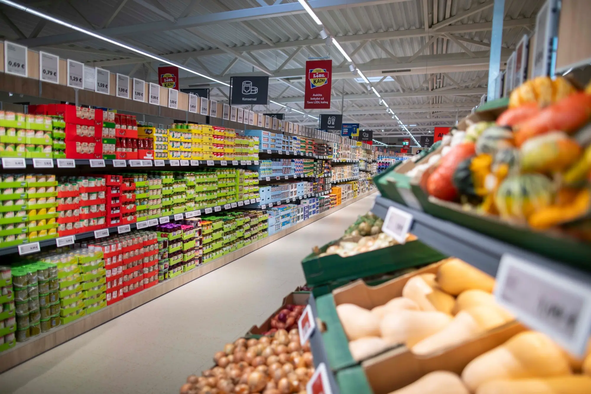 Interiér prodejny Lidl. Vlevo vidíme dlouhou řadu regálů plných trvanlivých potravin, jako jsou konzervy a balené výrobky, uspořádané podle barev a typů.