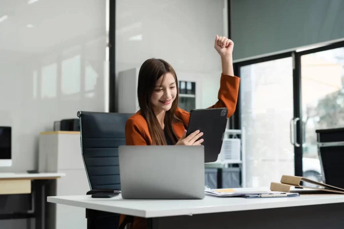 Mladá žena sedí v moderní kanceláři. Má na sobě oranžové sako a s úsměvem se dívá na tablet, který drží v jedné ruce.