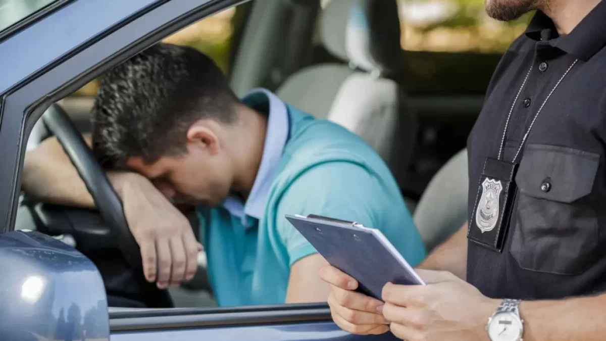 Policista komunikuje s řidičem sedícím v autě. Řidič má hlavu opřenou o volant a vypadá frustrovaně.