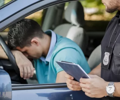 Policista komunikuje s řidičem sedícím v autě. Řidič má hlavu opřenou o volant a vypadá frustrovaně.