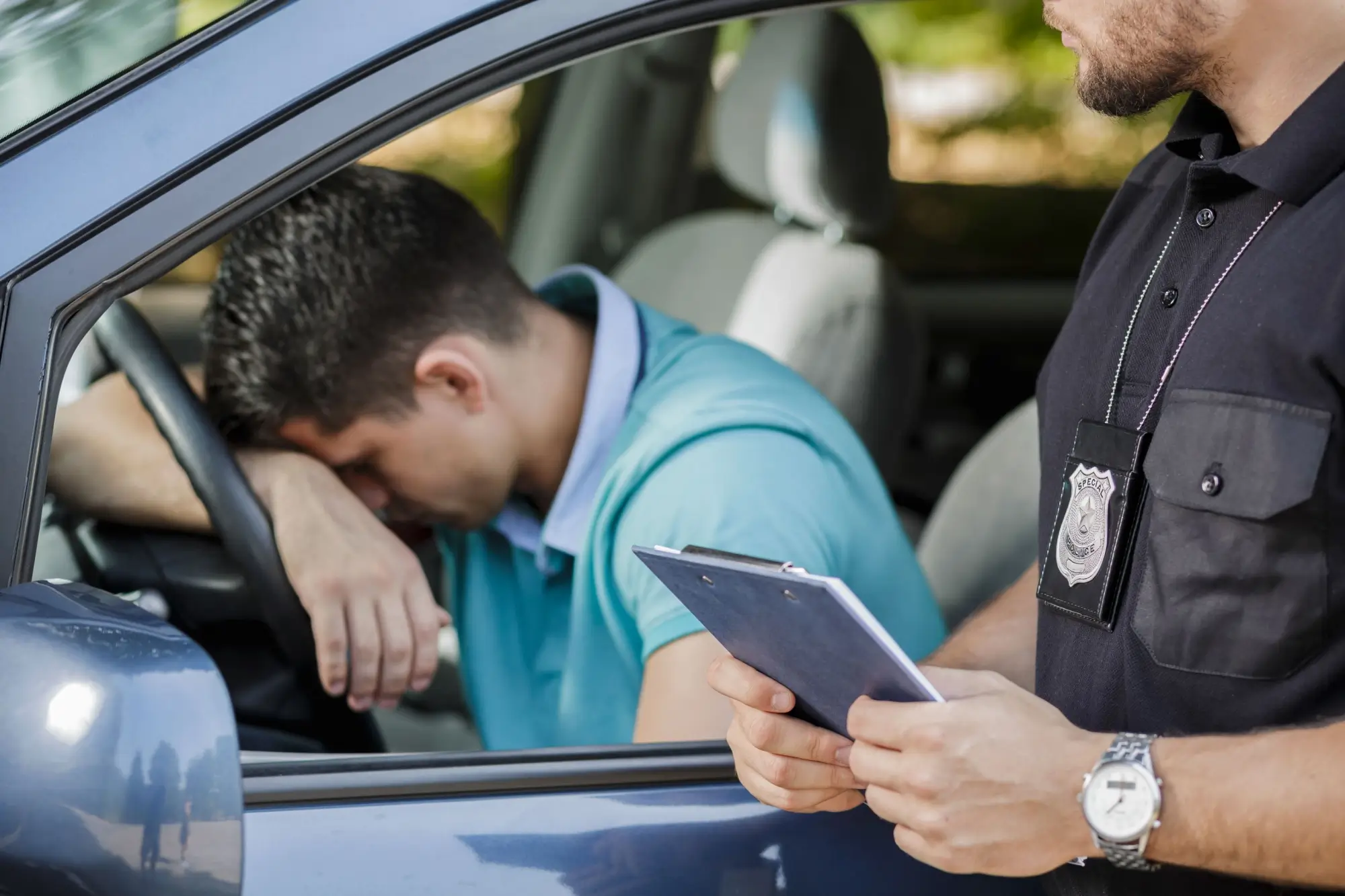 Policista komunikuje s řidičem sedícím v autě. Řidič má hlavu opřenou o volant a vypadá frustrovaně.