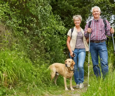 Starší pár na procházce v přírodě. Muž a žena kráčí po lesní cestě, mají batohy a nordic walkingové hole. Žena vede na vodítku světlého labradora. Všichni