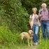 Starší pár na procházce v přírodě. Muž a žena kráčí po lesní cestě, mají batohy a nordic walkingové hole. Žena vede na vodítku světlého labradora. Všichni