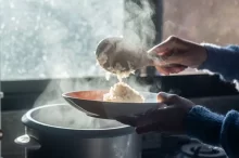 Žena nabírá čerstvě uvařenou rýži z hrnce pomocí dřevěné naběračky na talíř.
