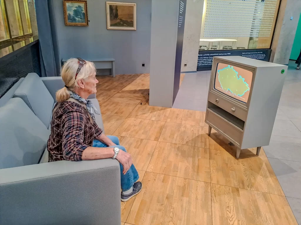 sen3 - Techsvět Starší žena sedí na sedačce v moderním interiéru, který imituje obývací pokoj z minulého století. Sleduje retro televizor, na jehož obrazovce je mapa Československa.