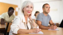 Starší žena sedící ve třídě u lavice, obklopená mladšími studenty. V ruce drží pero a zapisuje si poznámky