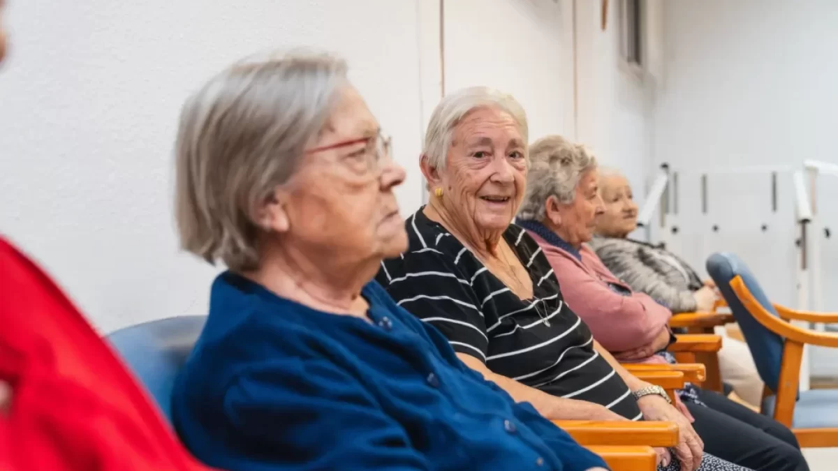 Skupina starších žen, sedí v řadě na židlích v interiéru, pravděpodobně v domově pro seniory, komunitním centru nebo čekárně. Všechny ženy působí klidně – jedna z nich se usmívá přímo do kamery, zatímco ostatní hledí stranou nebo před sebe.