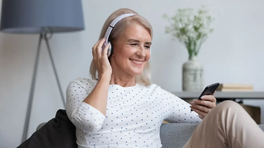 senior2 - Techsvět Usměvavá starší žena sedící pohodlně na gauči. Na uších má sluchátka a v ruce drží chytrý telefon, přes který poslouchá audiokurz.