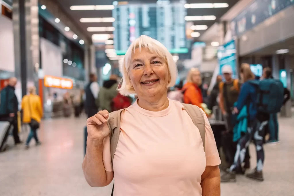 senior2 - Techsvět Seniorka s bílými vlasy, stojí ve veřejném prostoru – nejspíš na letišti nebo vlakovém nádraží. Na zádech má batoh a působí energicky a spokojeně