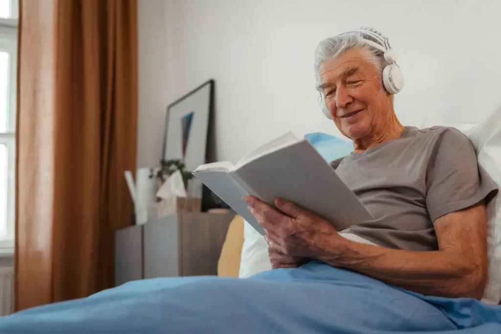 senior3 - Techsvět Starší muž ležící pohodlně v posteli. Na uších má sluchátka a v rukou drží otevřenou knihu, do které se soustředěně začítá.