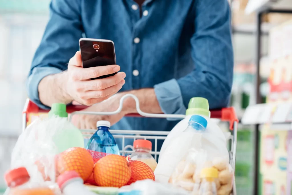 sleva3 - Techsvět Muž v obchodě, který drží chytrý telefon a opírá se o nákupní vozík plný potravin.