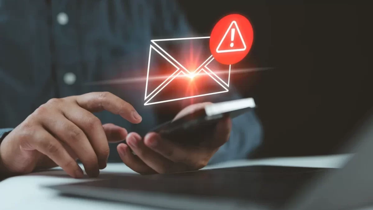 Muž drží chytrý telefon. Nad displejem je digitálně vložená ikona obálky s varovným vykřičníkem, což symbolizuje nebezpečnou nebo podezřelou zprávu.