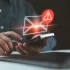 Muž drží chytrý telefon. Nad displejem je digitálně vložená ikona obálky s varovným vykřičníkem, což symbolizuje nebezpečnou nebo podezřelou zprávu.