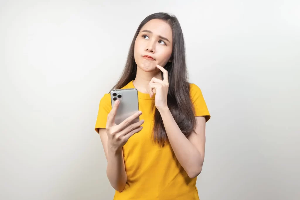 Mladá žena v žlutém tričku, která drží mobilní telefon a přemýšlí. 