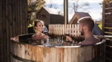 Muž a žena sedí ve venkovním dřevěném koupacím sudu, každý s vínem v ruce.